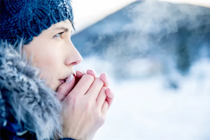 Девушка в вязаной шапке согревает дыханием замерзшие руки на фоне зимнего пейзажа